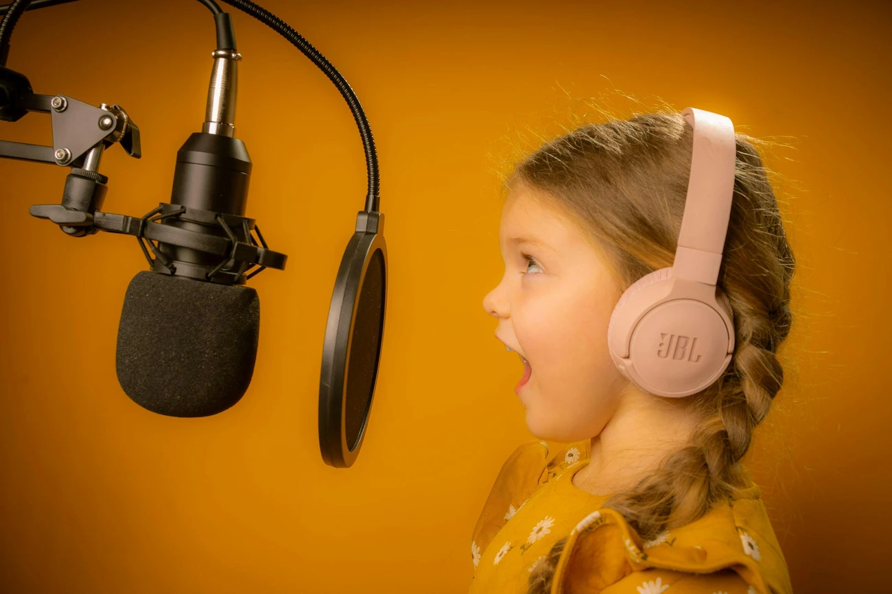 Criança loira cantando em um estúdio de música enquanto utiliza um headset rosa confortável exemplificando o melhor fone de ouvido infantil bluetooth.